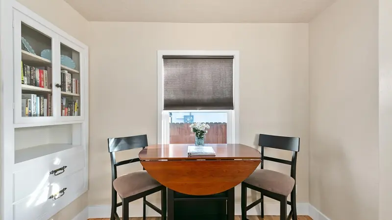 Small drop-leaf dining table with two chairs by a window, next to a built-in hutch with books.