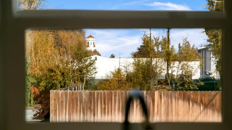 View through the kitchen window of trees, a wood fence and a clock tower in the distance.