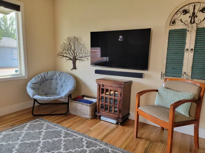 Wall-mounted TV above a small wooden cabinet, with a round chair, an armchair and a decorative shutter on the wall.